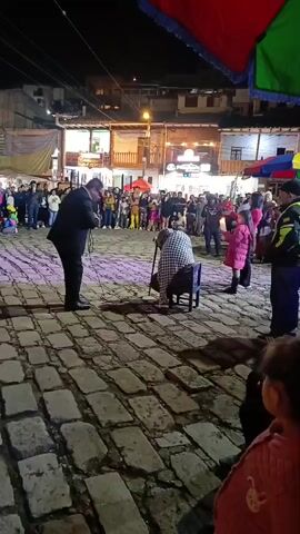 At night, a man in a black suit sings into a microphone in front of a crowd in a cobblestone plaza. An elderly woman sits on a chair nearby, leaning on a cane. The man gestures to the woman as he sings. People in the crowd are standing and watching the performance. Streetlights illuminate the plaza and buildings are visible in the background. There are colorful awnings overhead.