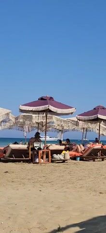 The video shows a beach scene with several people relaxing on lounge chairs under umbrellas. The umbrellas are purple with white fringe. The people are dressed in swimwear and casual clothing. Some are reading, some are talking, and others appear to be sunbathing. The beach is sandy, and the sea is visible in the background. A boat is also seen in the distance. The sky is clear and blue. The overall impression is one of a peaceful and relaxing day at the beach.