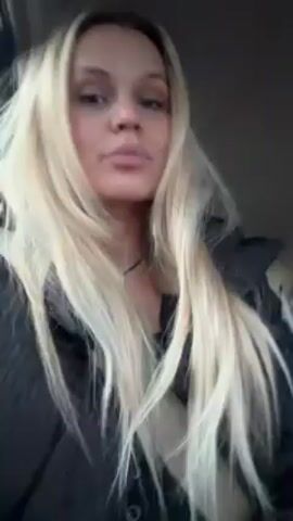 A woman with long blonde hair is seated in the passenger seat of a car. She is wearing a black jacket and a seatbelt. The camera is pointed at her face as she speaks and moves her head. The video then briefly shows the driver, a man with a beard, who is wearing a black turtleneck. The camera then returns to the woman, who continues to talk and move her head. The car appears to be moving.