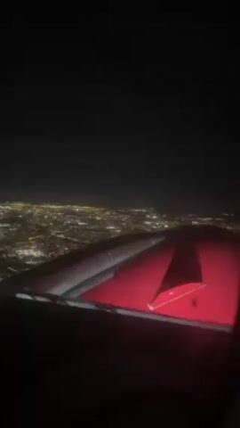 The video shows an airplane window view at night. The aircraft's wing is visible, illuminated with a red light. Below, a city sprawls out, sparkling with numerous lights spread across a vast area. The sky above is dark and clear.