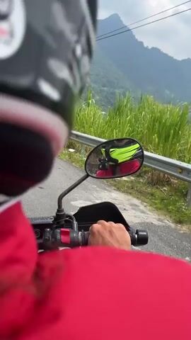 The video provides a first-person perspective of a motorcycle ride through a mountainous, rural landscape in Minh Sơn Commune, Vietnam. The rider is wearing a red jacket and a white helmet with red and yellow-green accents, with their hands visible on the motorcycle's handlebars.

The opening shot looks forward, with the rearview mirror reflecting the rider's helmet and the road behind. The surroundings feature lush green vegetation, including tall grass and trees, lining the side of the road. A silver guardrail runs alongside the paved road. In the background, green mountains rise under a cloudy sky, with power poles and lines stretching across the scene.

At the 0:01 mark, a yellow triangular road sign with a black border and a black squiggly arrow (indicating a winding road) becomes visible. The camera then shifts to show more of the expansive natural environment, highlighting the dense greenery and the distant mountain ranges. The prominent sound of wind suggests motion, indicating the motorcycle is in transit.