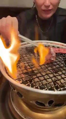 A person grills meat on a small, round grill placed on a table. The grill is filled with hot coals. The person uses tongs to turn the meat, which is emitting smoke. They appear excited and open their mouth in anticipation. The person is wearing a black shirt and jewelry. The environment seems to be a restaurant setting.