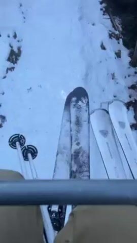 The video shows a view from a ski lift ascending a snow-covered slope. The person filming is holding ski poles and has skis resting on the footrest of the ski lift. The slope is bordered by trees, and other ski lift chairs are visible in the distance. The snow cover is uneven with patches of exposed ground and rocks. The weather appears to be sunny.