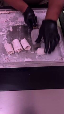 A person wearing black gloves uses a rectangular metal spatula to scrape ice cream on a metal surface. The surface appears frozen. Four ice cream rolls sit on the surface. The rolls are white with small bits of brown or pink inside. The person slides the spatula across the metal surface. The surface of the area is covered in a layer of white ice cream. The machine appears to be black. A pink light shines on the metal surface.