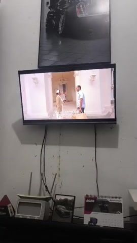 A medium-shot video shows a wall-mounted flat-screen television displaying a scene from what appears to be a television program. Two men are visible on the screen. One man, dressed in traditional Islamic attire, stands facing the other. The second man, wearing a white shirt and blue pants, is holding a bag and standing to the right of the first man. They appear to be in an indoor setting with white walls and a doorway in the background. Below the television, on a shelf, are several items including a framed photograph, a calendar with the year 2026 visible, a small red pyramid-shaped object, and a white electronic device. The wall behind the television has a large framed picture of a black car. Black cables are visible hanging from the back of the television.