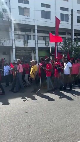A group of people march down a street, carrying red flags. Most of them wear red shirts and some have red hats. The march takes place in front of a multi-story white building. Some people are wearing other colors, such as yellow, blue, and white. A bus passes in the foreground.
