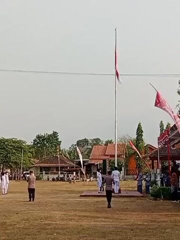 A group of people are gathered on a field for a flag raising ceremony. A tall flagpole stands in the center of the frame, with the Indonesian flag (red and white) partially raised. Several individuals in uniform are standing around the flagpole, and others are observing from a distance. There are buildings and trees in the background. The sky is overcast.