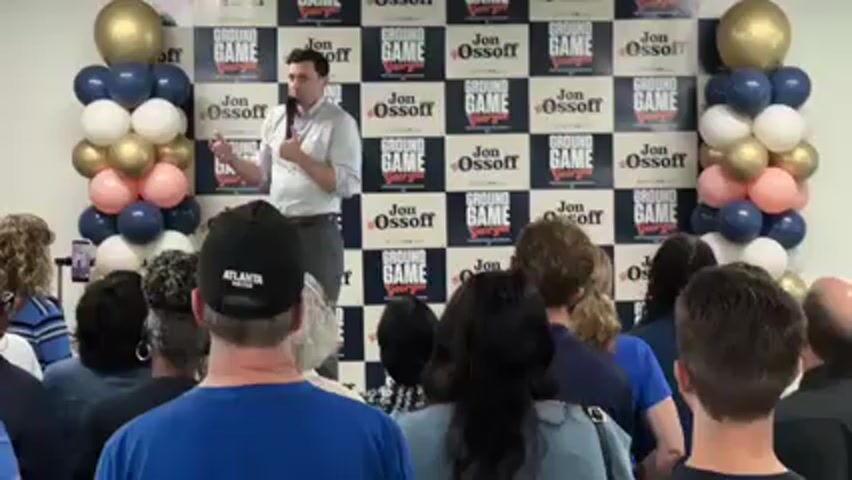 In an indoor setting, a man, identified as Jon Ossoff, stands on a slightly raised platform, addressing a seated audience. He holds a microphone in his left hand and gestures with his right as he speaks. Ossoff is dressed in a light-colored button-down shirt and grey trousers.

Behind him, a patterned backdrop features alternating squares of "GROUND GAME Georgia" and "Jon Ossoff." Flanking the backdrop on both sides are decorative balloon columns in colors of gold, navy blue, white, and peach.

The audience, composed of diverse individuals, faces the speaker. Most are seated, and some are looking directly at Ossoff, while others are seen from behind. One person in the foreground wears a black baseball cap with "ATLANTA UNITED" written on it. To the left, a person holds up a phone, possibly recording the event.

The scene appears to be a campaign event or town hall meeting in Decatur, United States, given the political branding and the speaker's engagement with the crowd.