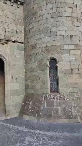 The video shows a view of an arched stone gateway with people walking through it. A young girl in a white hat, blue shorts, and pink sneakers stands in the foreground on the left, holding a bag or bundle. To the right of the gateway, a person in a green outfit stands and appears to be taking a photo with a phone. Beyond the archway, other people are visible walking down a cobblestone path lined with stone buildings. Lush green vines climb the stone wall on the left side of the gateway. The overall environment suggests an old, possibly historic, stone structure.