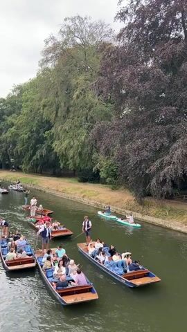 A video shows multiple boats, some with passengers, navigating a river. A large, old, stone building with multiple windows and chimneys is visible on the left bank. Trees line the opposite bank. Several people are seated in the boats, while others stand and appear to be steering them. Some of the boats are being punted, a method of propulsion using a long pole. On the left bank, there is an outdoor seating area with tables and umbrellas, and people are seated at the tables. The sky is overcast. The video pans slightly to the right, revealing more of the river and the building.