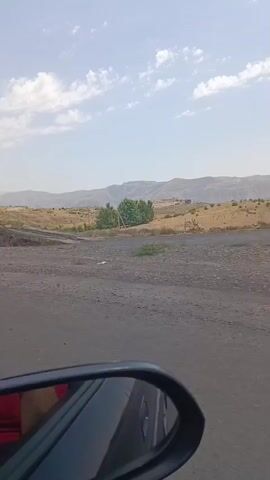 The video appears to be shot from inside a vehicle, looking out the passenger side window. The road surface is rough and appears to be dirt or gravel. In the foreground, the side mirror of the vehicle is visible. The landscape is arid and hilly, with sparse vegetation consisting of dry grass and some green bushes. In the distance, there are rolling hills and mountains under a bright, partly cloudy sky. Two traffic signs are visible. The first sign, near the beginning of the video, is a triangular warning sign with a black symbol depicting a road that forks into three paths, with the main road continuing straight and a smaller road branching off to the right. The second sign, further along the road, is a rectangular sign with red and white diagonal stripes, indicating a curve in the road, likely a sharp one to the left. There is a visible bridge or overpass in the mid-ground, and some scattered debris, including a white plastic bag, on the side of the road. No people are visible in the video. The overall impression is of driving through a rural, undeveloped area.