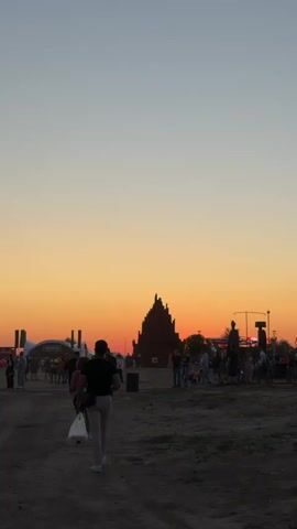 The video opens with a wide shot of an outdoor festival at dusk. The sky is a gradient of orange and pale blue. In the foreground, a man walks away from the camera, carrying a white plastic bag in his left hand. His back is to the viewer, and he is wearing a dark t-shirt and light-colored shorts. Behind him, several other people are visible, some walking and some standing. A large, dark, pyramid-shaped structure with jagged edges is prominent in the mid-ground. Further back, a white stage or tent structure with a sign is visible, along with more people and what appear to be temporary fences and lighting rigs. The ground is dirt and appears somewhat uneven. The overall atmosphere suggests a gathering or event taking place as the day ends. As the video progresses, the camera pans slightly and zooms in on different areas of the festival grounds, showing more details of the crowd and structures. The lighting suggests the sun has recently set or is very low on the horizon. There are no overt interactions between individuals that are clearly depicted, but the presence of numerous people suggests a social gathering. The environment is clearly an open, outdoor space designed for a large event.