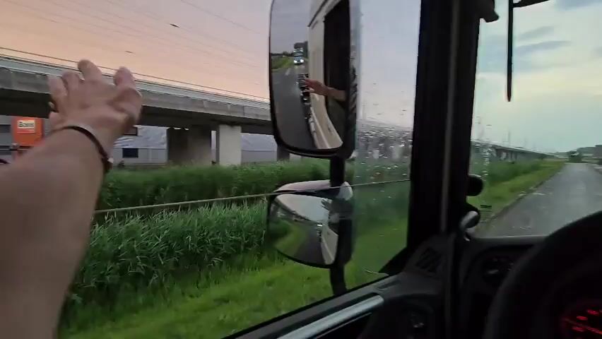 A person's left arm is visible in the foreground, extending towards the left and slightly upward, with their hand raised as if waving. The arm is positioned near the side mirror of a truck cab. The side mirror reflects a view of the road behind the truck, with another vehicle visible in the distance and a person inside that vehicle also waving. Raindrops are visible on the truck's window and side mirror, suggesting wet weather. In the background, there is a raised overpass or bridge structure, with greenery and vegetation on the side of the road. The sky appears overcast.
