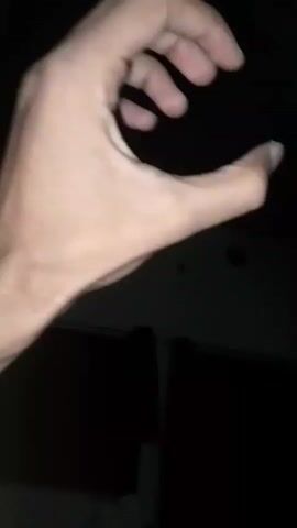 The video displays a close-up of a person's left hand against a dark background. The hand is illuminated, revealing skin tone, veins, and fingernails. The fingers are in motion, transitioning from a partially closed position to a fully closed fist, then opening to a C-shape. A small, metallic object, possibly a screw or a bead, is visible between the thumb and index finger at one point. The background is dark and indistinct, with faint shapes suggesting indoor curtains or walls and a ceiling fixture. The lighting is dim, with light sources appearing to come from above and slightly to the side of the hand.