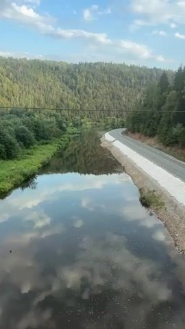 This video shows a view from a moving vehicle, likely a train, looking out at a winding river surrounded by lush green forested hills. The sky is partly cloudy with a blue sky visible. The river reflects the sky and the trees on its surface. A road is visible on the right side of the river, with a white gravel path alongside it. The vegetation appears dense and varied, with deciduous and coniferous trees. The camera pans slightly to the right, revealing more of the forested landscape and the curve of the road.