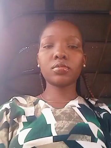 The video is a close-up shot of a woman's face. She has brown skin, dark hair pulled back in braids, and is wearing a top with a geometric pattern in green, white, and brown. She also has a thin gold necklace and small pearl earrings. The background appears to be the interior of a vehicle, possibly a rickshaw, as the exterior is visible through the windows on either side of her. The light is bright, and the video seems to have been taken during the day. She looks directly at the camera with a neutral expression. There is some movement visible in the background.