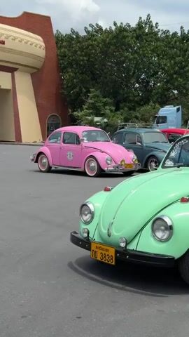 The video shows a line of vintage Volkswagen Beetles parked in front of a large, arched building that appears to be a border gate. The building is cream-colored with a red archway and features text in Vietnamese. The Beetles are in various colors, including mint green, pink, teal, red, and blue. The mint green Beetle in the foreground has luggage on its roof rack. A person in a green shirt is visible inside the mint green Beetle. The pink Beetle has a decal on its side. The background consists of trees and a cloudy sky. There is also a shipping container with Chinese text visible behind the Beetles. The overall scene suggests a travel or event related to vintage cars at an international border crossing.