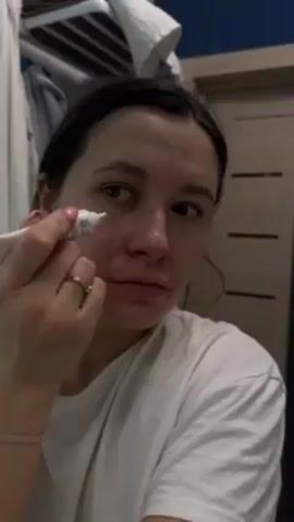 A fair-skinned woman with brown hair pulled back from her face is shown in a close-up shot applying cream around her eyes from a small white tube. She is wearing a white t-shirt and a gold ring. The bathroom setting includes white walls, a door, and a towel rack with towels.