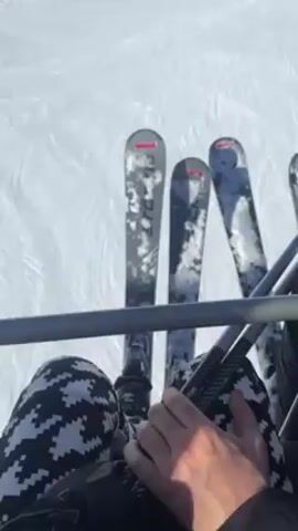 The video shows a first-person view from a ski lift, looking down at skis and then up at a snow-covered ski slope with other skiers and ski lifts. The person filming is wearing patterned black and white pants. The skis are also white with grey and black patterns and the words "DC" are visible on the top. The snow is white and marked with tracks. The sky is blue and clear. The ski lift is moving up the slope.