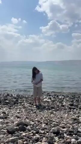 A woman stands on a rocky beach next to a calm body of water under a partly cloudy sky. She is wearing a white top and tan shorts. She moves her hair away from her face and then stands still, looking toward the camera. The water is a light turquoise color, and there is land visible in the distance across the water. The beach is made up of dark-colored rocks.