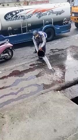 A man in a white and blue vest, dark pants, and a blue cap is pouring liquid from a black bucket onto a wet street. He is standing next to a large white tanker truck with blue accents and text that reads "SIBOLANGIT 2 TgN SUANGGI". A pink scooter is parked on the left side of the frame. The street surface appears to have dark, oily stains and is covered in puddles. The man pours the contents of the bucket onto the oily substance and then walks away from the camera, toward the tanker truck.