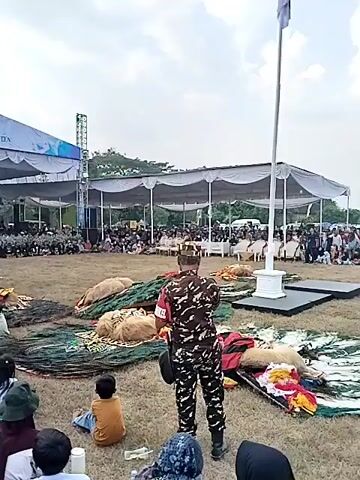 The video shows an outdoor performance area. In the background, a large tent with a banner reading "41st ANNIVERSARY PONDOK PESANTREN NURUL HUDA" is set up, with a stage and screen beneath it. A crowd of people, many wearing dark clothing, are seated in front of the stage. In the foreground, a grassy area is visible, with more people sitting and watching. Several large, bundled objects, possibly costumes or props, are scattered on the ground. A performer in traditional attire is actively dancing or moving in the center of the performance space. A person in a camouflage uniform stands in the foreground, facing the performance area. The sky is overcast.