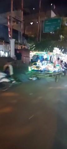The video shows a nighttime street scene.  The camera is moving, suggesting it is being carried or is on a vehicle. In the foreground, the ground is visible, appearing to be a paved surface.  To the left, a white structure, possibly a pillar or wall, is present.  Further into the scene, a series of illuminated carts or small rides are lined up. These rides are decorated with colorful lights, and one appears to be a child's ride shaped like a motorbike.  Several people are visible near these rides, some appearing to be children, and others adults. In the background, buildings and trees are dimly lit. Several motorcycles are parked along the side of the street. The overall impression is of a lively night market or fairground setting.