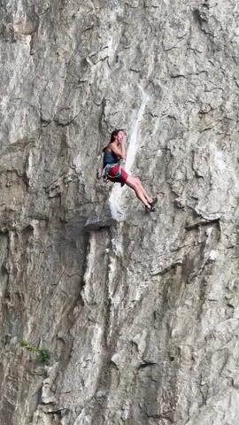 A rock climber is ascending a sheer rock face. The climber, wearing a dark tank top and red shorts, is tethered to a rope that runs vertically up the cliff. Their body is angled against the rock as they reach for a higher handhold. The rock face itself is rugged and textured, with visible cracks and ledges. The surrounding environment is not clearly visible, but the focus is on the climber's ascent and the imposing rock wall. The video footage is slightly shaky at times, indicating it may have been captured by a handheld device.