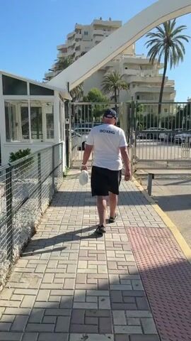 A man in a white t-shirt with “BOXING” printed on the back and a blue baseball cap is walking on a brick sidewalk. He is wearing black shorts and sandals. He is carrying a white plastic bag in his left hand. He is walking towards an open metal gate, leading to a street with parked cars and palm trees. The environment appears sunny and possibly coastal.