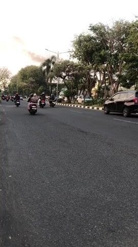 A road view shows traffic with several motorcycles and a black car. The sky is overcast, and there is smoke in the distance. The road is paved, and trees line the right side of the road. Billboards and power lines are visible on the left. The motorcycles are moving in the same direction as the car.