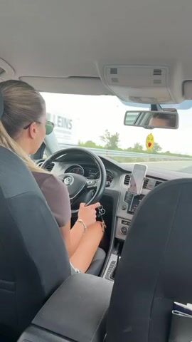 The video shows the interior of a car from the perspective of a rear passenger. A woman is driving, with her back to the camera, wearing sunglasses and a dark top. Her hands are on the steering wheel, and she is holding keys. The car is on a highway, with trees and fields visible outside. The car's dashboard and infotainment system are visible. A phone is mounted on the dashboard and appears to be displaying navigation. A yellow air freshener is hanging from the rearview mirror. The overall scene suggests a daytime road trip.