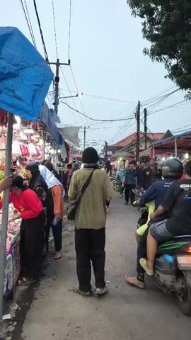A vertical, full shot captures a crowded street market under an overcast sky. On the left, a stall draped with a blue tarp displays various items, possibly clothing, with several people, including women in headscarves and a woman in a red top, browsing. In the center, a person wearing a green shirt and black pants walks away from the camera, carrying a bag over their shoulder. To the right, the street is busy with activity. A motorcycle carrying two people is moving away from the viewer. Several other individuals are walking or standing along the right side of the street, where more stalls are visible, suggesting a commercial area. Power lines are strung across the upper portion of the frame, and trees are visible in the background on the right. The overall scene depicts a lively and bustling market environment.