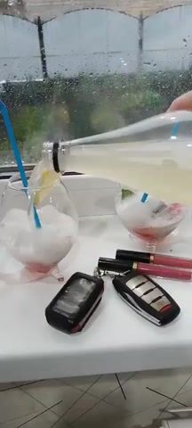 This is a close-up, first-person perspective video of a person pouring a yellow liquid from a clear bottle into two glasses. The glasses contain ice and some fruit, possibly lime and strawberry, along with blue straws. One glass is filled with a white, frothy mixture and garnished with a lemon slice. The other glass has a pinkish hue.

In the foreground, on a white surface, are two car keys and two tubes of liquid lipstick, one black and one pink. The background shows a window with rain streaks, suggesting it is daytime and raining. The scene appears to be indoors, possibly on a windowsill, with greenery and indistinct structures visible through the rain-streaked glass. The overall mood is relaxed and perhaps a bit melancholic due to the rainy weather, contrasted with the refreshing drinks and makeup. There is no visible text. The context provided indicates the location is Novostroika, Russia.