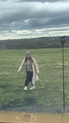 A woman in a tan trench coat, gray sweater, black pants, white sneakers, and a gray beanie walks towards a phone on a tripod in a grassy field. The sky is overcast. Trees and houses are visible in the background. The person filming is likely in a car or behind a window.