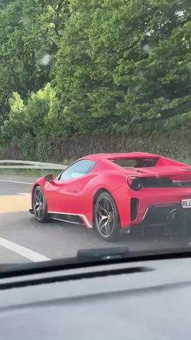 A red Ferrari 488 Pista is driving on a road, filmed from another vehicle. The car is on the right side of the road and the video vehicle is behind it. The Ferrari has a license plate that starts with "LU." The environment is sunny with some trees alongside the road and buildings in the background. There are also highway signs visible.