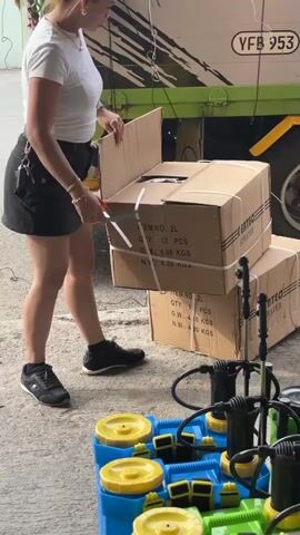 A person is cutting open a cardboard box with scissors. The box is stacked on top of another identical box. The boxes have labels that read "ITEM NO.: 2L", "QTY.: 12 PCS", "G.W.: 4.80 KGS", and "N.W.: 4.20 KGS". The person is wearing a white t-shirt, a black skirt, and black shoes. In the foreground, there are several blue and green plastic containers with yellow lids, and black spray nozzles and hoses. In the background, there is a truck with green and white signage. The scene appears to be outdoors on a concrete surface.