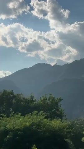 The video shows a moving shot of a mountainous landscape under a partly cloudy sky. In the foreground, lush green trees and foliage are visible. The middle ground features a green field, possibly farmland. In the background, the hazy outline of rolling mountains dominates the scene. A small, distant structure, possibly a tower or a building, is visible on one of the mountain peaks. The lighting suggests it is daytime, with sunlight breaking through the clouds. The camera appears to be in motion, possibly from a vehicle, as the perspective shifts slightly throughout the short clip.