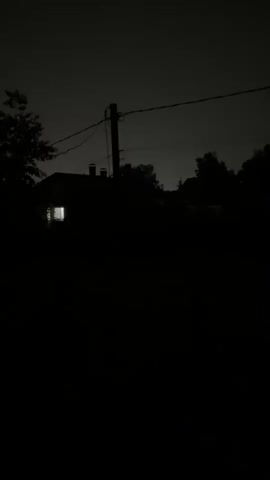 A short video clip shows a dark, outdoor scene at night. In the middle ground, a silhouette of a building with a lit window is visible among trees. A utility pole with power lines stretches across the frame. The foreground and background are predominantly black, indicating a very dark environment with minimal light. The clip is approximately 4 seconds long.