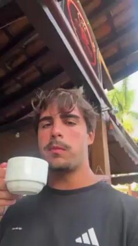A young man with light brown hair and a mustache is drinking from a white ceramic cup. He is wearing a black t-shirt with an Adidas logo. The background shows a partially visible wooden roof structure, with some foliage and a sign in the distance. The camera is positioned at a low angle, looking up at the man.