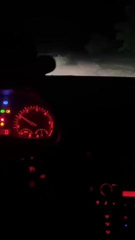 The video shows the interior of a car at night. The dashboard lights are red, illuminating the speedometer, tachometer, and various indicator lights. The headlights are on, casting a beam of light onto a dirt road ahead, with trees and brush visible on either side. The car appears to be in motion.