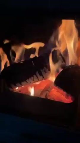 The video shows a close-up view of a fire burning inside a wood-burning stove or fireplace. There are logs inside, some glowing red with heat, and flames are dancing around them. The fire is the main source of light in the dim environment. The interior of the stove appears dark, and the edges of the stove frame are visible.