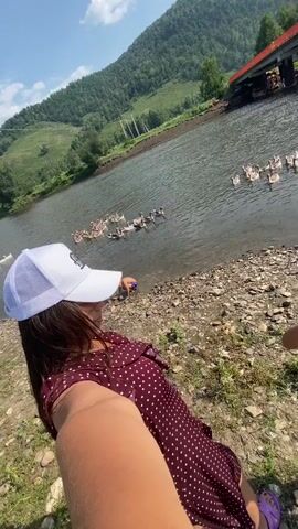 A woman in a white baseball cap and a maroon polka dot dress is taking a selfie on a rocky shore next to a body of water. Geese are swimming in the water. A bridge is visible in the background, as well as a forested mountain. A child is briefly visible in the frame.