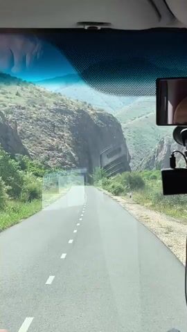 A dashcam view shows a paved road winding through a mountainous landscape. The road has a dashed white line down the center. The mountains on either side are covered in dry vegetation and rocks. Trees and bushes grow along the sides of the road. A cell phone is mounted to the windshield on the right side, and a dashcam is also visible.