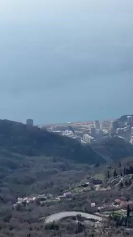 The video shows a high-angle view of a coastal city nestled among hills and mountains, transitioning to reveal a clear blue sea with two small islands in the distance. The city consists of a mix of high-rise buildings and smaller structures, scattered among dense vegetation. The hills in the foreground are covered in trees that are mostly leafless, suggesting it may be early spring or late fall. There are also scattered red-roofed buildings visible on the slopes. The sea is calm, with a light-colored haze obscuring the horizon. The overall impression is one of a scenic view from a high vantage point.