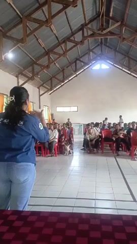 The video shows an indoor event with an audience seated on red chairs. A woman in a blue shirt and jeans stands at the front, facing the audience. She appears to be speaking or presenting. Two dogs are visible among the seated attendees. The room has a high, sloped ceiling with exposed wooden beams and a corrugated metal roof. The walls are light-colored, and there are windows along one side. The floor is tiled. The overall atmosphere seems to be a community gathering or educational session.