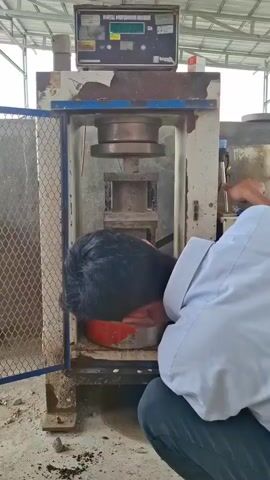 A person in a light-colored collared shirt and dark pants is crouched down in front of a large, industrial testing machine. Their head is bowed and positioned directly over a cylindrical object, possibly a concrete sample, resting on the machine's lower platen. The machine has a digital display that reads "0.5" and then changes to "0." The machine has a metal cage door open on the left side. The environment appears to be an indoor workshop or laboratory with a concrete floor and a corrugated metal roof overhead. Debris is scattered on the floor around the machine.