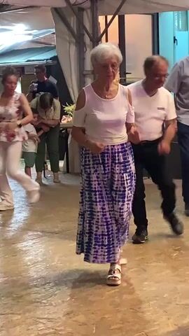 A group of people are dancing outdoors at an event. In the foreground, an elderly woman with short gray hair is wearing a white off-the-shoulder top and a long blue and white tie-dye skirt. She is dancing with her arms extended and appears to be smiling. Behind her, an older man in a white t-shirt and black pants is also dancing, as is another older man in a light blue button-down shirt and dark pants. In the background, several other people, including a child, are visible, standing or observing the dancers. The environment appears to be a temporary outdoor venue, possibly a festival or street fair, with a tented roof and some tables visible. The ground is paved.