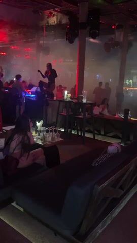 The video shows a nightclub or bar with a lively atmosphere. Red lighting and haze fill the air. A DJ stands on a raised platform, and people are dancing. Several customers are seated at tables and on lounge furniture, some with drinks. Waitstaff move among the tables, serving customers. The camera pans across the scene, showing different areas of the club. The overall impression is that the venue is crowded and energetic.