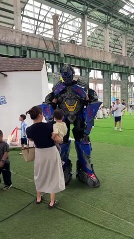 A woman is holding a child and taking a picture of a person in a full Optimus Prime costume on a green turf floor indoors. The costume is blue, black, and red with yellow accents. The woman is wearing a dark top and a light-colored skirt. The child is wearing a light-colored shirt. The background shows an industrial-looking building with exposed beams and a pitched roof. Other people are visible in the distance, including a man in a white t-shirt and shorts walking on the turf. There are some white structures and a sign with Chinese characters visible to the left of the robot.