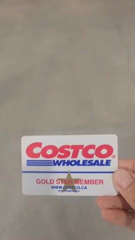 This is a first-person perspective video clip, likely taken within a large retail store. The floor is concrete and shows some wear, including cracks. In the foreground, a Costco membership card is held up, with the "Costco" logo and "Gold Star" visible.

In the mid-ground, several people are present. One person, wearing a colorful, flowing dress and white sneakers, is pushing a shopping cart and walking away from the camera. Another person, wearing a red vest and jeans, stands near a scanner for membership cards, holding what appears to be a newspaper or magazine. Behind them, a large trash bin with a plastic liner is visible. Further in the background, more shoppers and merchandise displays can be seen, including large screens displaying imagery. The lighting suggests it is daytime, and the overall impression is that of a busy shopping environment, likely a Costco store in Mississauga, Canada, given the context. The camera angle and focus suggest the user is about to scan their membership card to enter the shopping area.